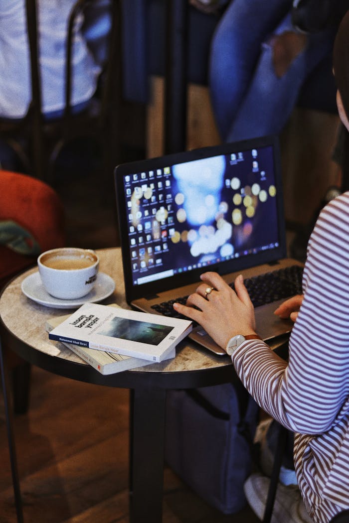 Crop anonymous female remote employee working on netbook at cafeteria table with books and cappuccino against people